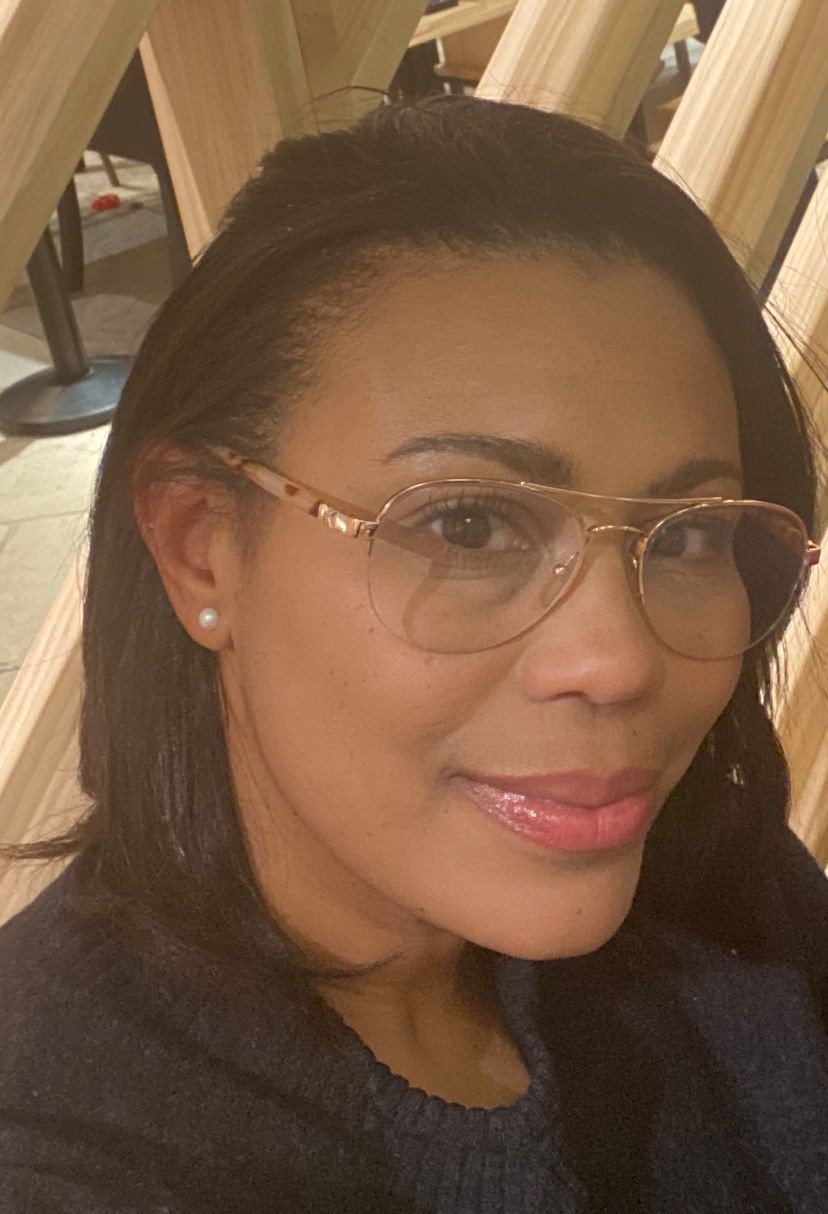 Smiling person wearing glasses, sitting indoors with a wooden structure in the background.