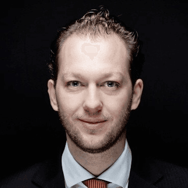 Man in a suit and tie with a dark background, facing the camera with a neutral expression.